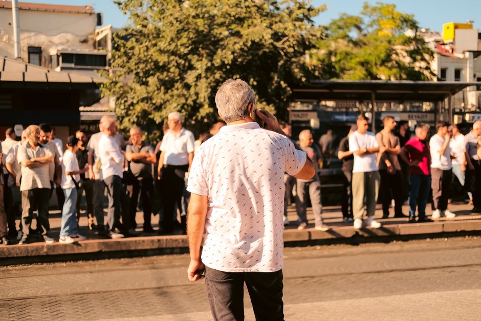 A busy urban bus stop with a crowd of people waiting, captured in warm sunlight.
